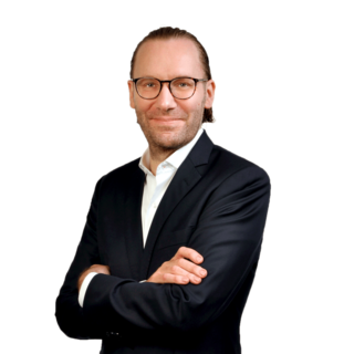 Mann mit Brille im dunklen Anzug, lächelnd mit verschränkten Armen vor schwarzem Hintergrund